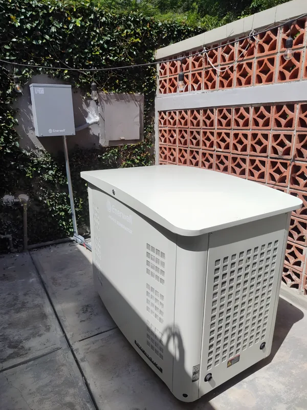 Planta estacionaria Enerwell instalada junto a un muro de celosía de barro, con su tablero de transferencia automática en la pared