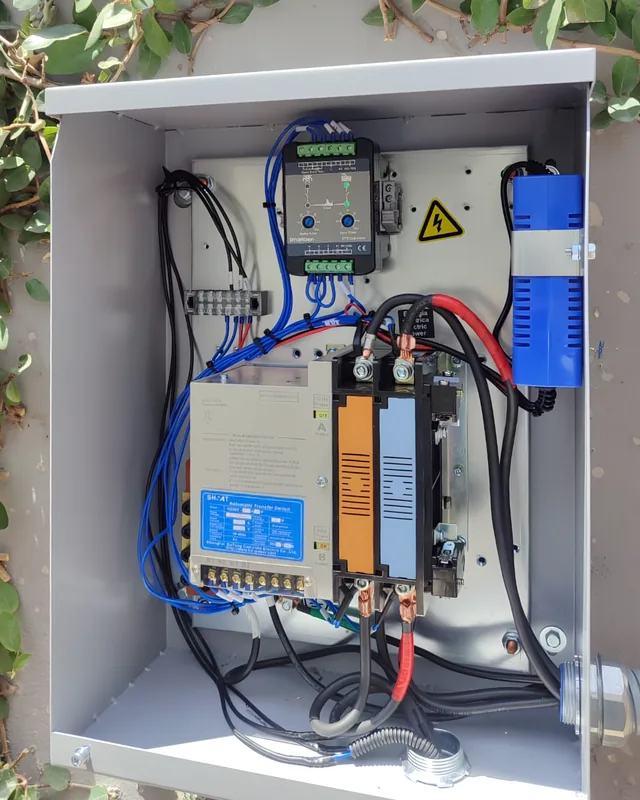 Tablero de transferencia automática abierto con su controlador y conmutador, montado en un muro con enredadera